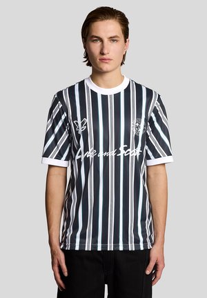 Giovane uomo con capelli corti castani che indossa una maglietta a maniche corte a righe verticali bianche e nere con colletto bianco, in piedi davanti a uno sfondo uniforme.