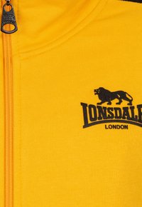 Žltá mikina na zips vyrobená z mäkkej textílie. Na prednej strane sa nachádza čierne logo levu a text "Lonsdale London". Textúrovaný povrch.