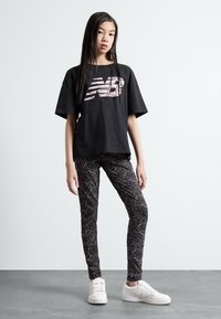 Crna majica kratkih rukava s ružičastim New Balance logotipom, u kombinaciji sa prilagođenim crnim tajicama s uzorkom valovite linije, bijelim tenisicama.