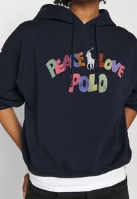 Marinblå hoodie i bomullsblandning, med en flerfärgad grafik som säger "PEACE LOVE POLO" och en vit polo-spelare-logotyp.