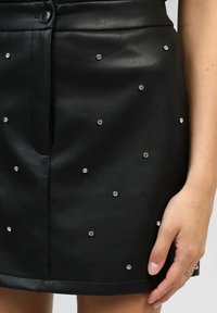 Jupe en cuir noir avec de petites embellissements en strass éparpillés, fermeture à boutons à la taille, et une main reposant à côté avec des bagues.