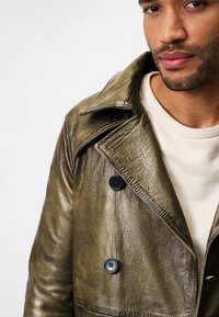 Olive green leather trench coat with a crinkled texture, large collar, and black buttons. Worn over a light cream sweater.