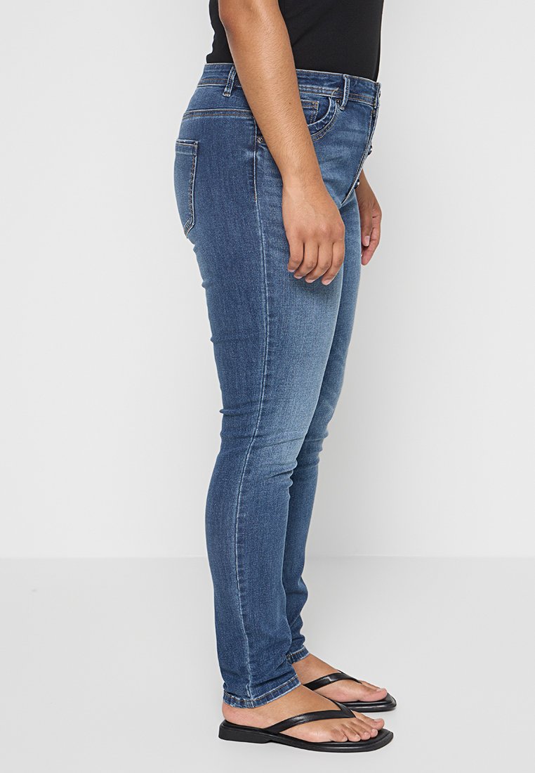 Schmale blaue Denim-Jeans mit einem ausgewaschenen Finish, fünf Taschen, Gürtelschlaufen und einer mittelhohen Taille. Getragen mit schwarzen Flip-Flops.