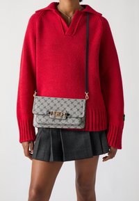 Persona che indossa un maglione rosso a maglia, una gonna nera plissettata e una borsa a tracolla grigia con motivo e dettagli in oro sulla fibbia.