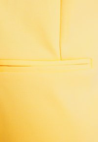 Détail de poche en tissu jaune avec une finition texturée ; présente un design à coupe droite et un accent de couture horizontale en haut.