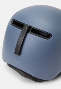 Sivoblu čelada z mat površino, s črnim prezračevalnim trakom in nastavljivim trakovi. Besedilo "TAGGERT" vidno na dnu.