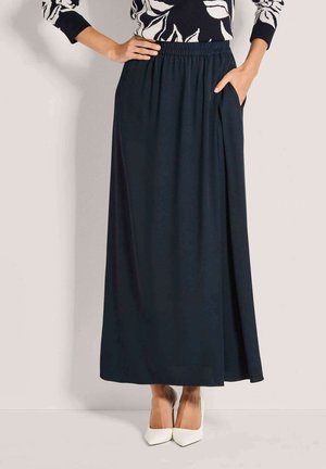 Femme portant une longue jupe noire à taille élastique avec poches, associée à des escarpins blancs à bouts pointus et un haut à motif noir et blanc.