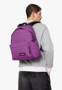 Mochila morada con un gran compartimento principal, bolsillo frontal y detalles en cremallera negra. Hecha de tela duradera, cuenta con correas de hombro ajustables.