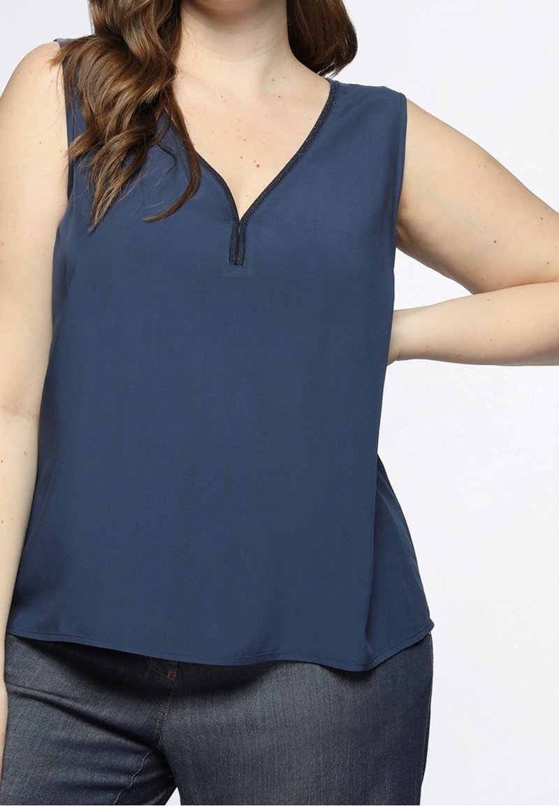 Femme portant un top sans manches bleu marine avec un col en V et un jean foncé, posant avec une main sur la hanche devant un fond clair uni.