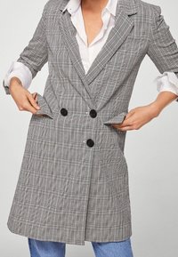 Blazer doppiopetto grigio a quadri con bottoni neri, revers a punta e tasche laterali. Tessuto liscio con vestibilità sartoriale.