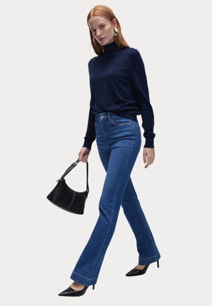 Donkerblauwe coltrui, aansluitende blauwe flared jeans, zwarte handtas met strap en zwarte puntige hakken, gepresenteerd in een looppose.