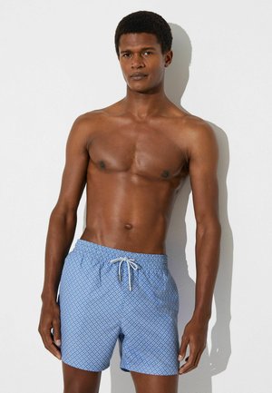 Skjorteløs mand med kort krøllet hår iført blå badeshorts med hvid snøre, stående op ad en hvid væg.