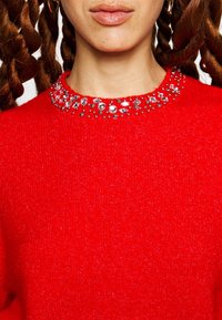 Nainen, jolla on kierteiset letit ja joka on pukeutunut kirkkaan punaiseen villapaitaan, jonka kauluksessa on jalokiviä, sekä kultaiset renkaat korvissa. Huulten yläosa näkyy osittain.