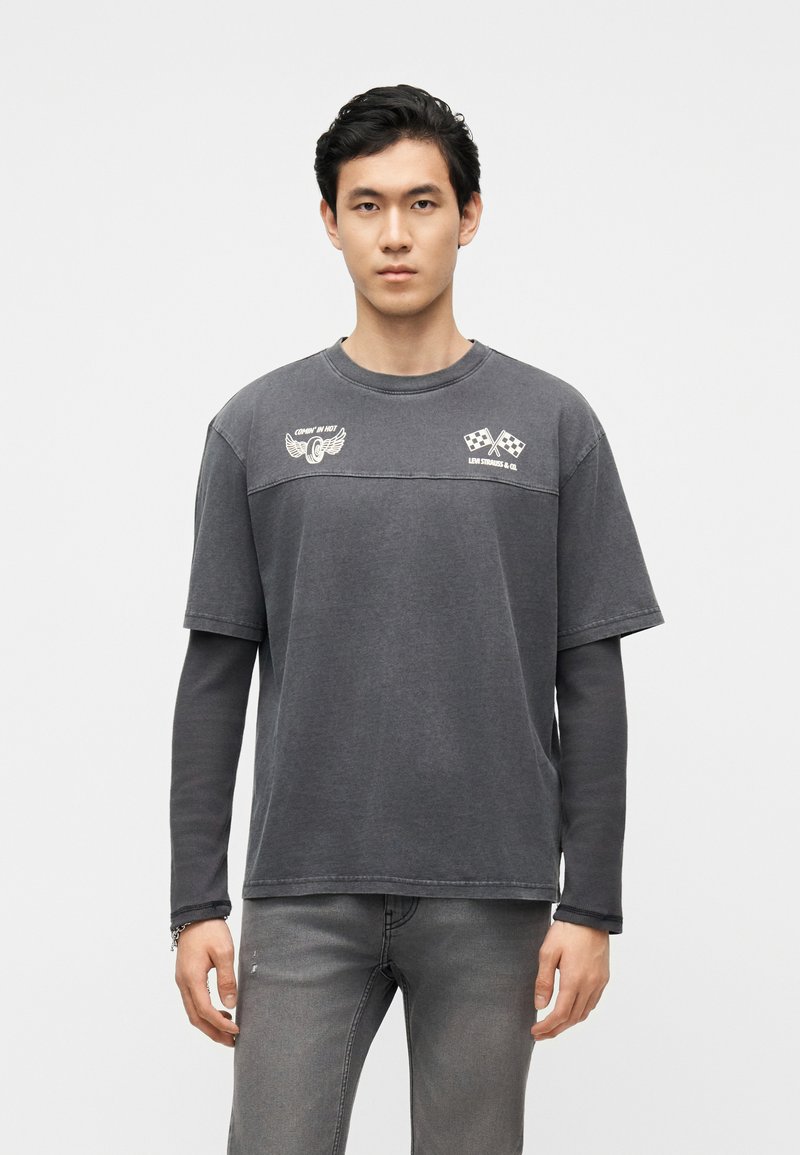 Young man wearing layered dark gray long-sleeve shirt and T-shirt with white racing and wing logos, standing against plain light background.