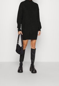 Robe pull noire en maille avec col haut, manches longues et fentes latérales. Portée avec des bottes noires montantes jusqu'aux genoux et un petit sac noir à bandoulière.