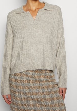 Femme portant un pull côtelé beige avec col et une jupe à carreaux gris et marron, debout devant un fond uni.