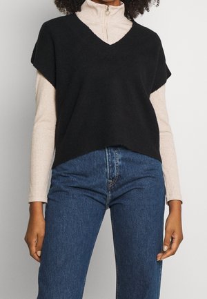 Femme portant un gilet-pull noir à manches courtes en V par-dessus un col roulé côtelé beige à fermeture éclair et un jean bleu taille haute.