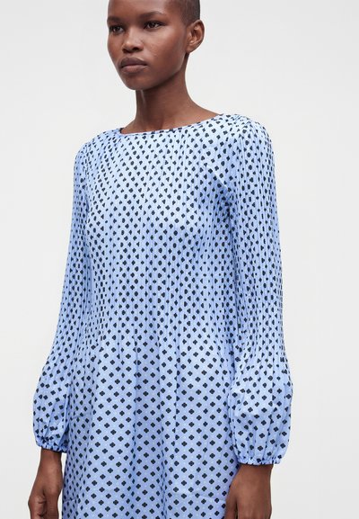 Femme portant une robe bleu clair à manches longues avec de petits motifs floraux noirs et des poignets élastiques froncés, sur un fond clair uni.