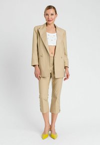 Blazer beige su misura e pantaloni a vita alta, abbinati a una blusa bianca e ballerine gialle a punta. Tessuto morbido con dettagli a bottoni sui pantaloni.