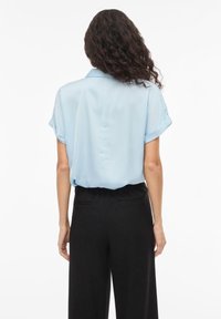 Ανοιχτό μπλε μπλούζα με κοντά μανίκια και απαλή υφή, με γυρισμένα μανίκια και λεπτομέρεια με κουμπιά στην πλάτη, συνδυασμένη με μαύρο ψηλόμεσο παντελόνι.