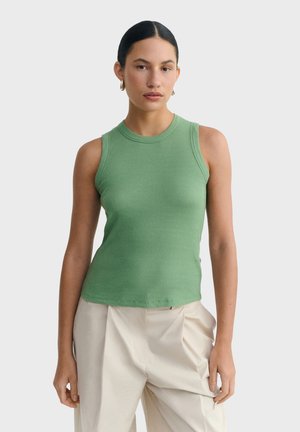 Femme aux cheveux foncés portant un haut sans manches vert côtelé et un pantalon beige plissé, debout devant un fond clair uni.