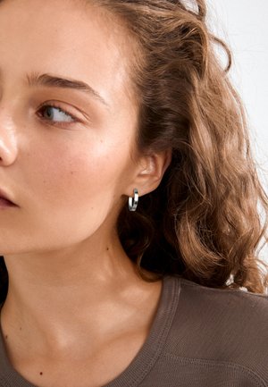 Žena s vlnitými hnedými vlasmi, ktorá nosí malé strieborné kruhové náušnice a hnedý top, pozerá sa doľava na jednoduchom svetlom pozadí.