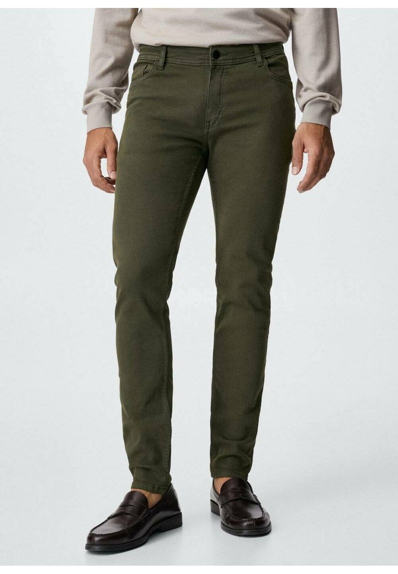 Homme portant un pantalon slim vert olive, une chemise beige à manches longues et des mocassins en cuir marron foncé, debout sur un fond clair.