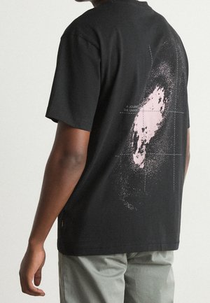 Osoba w czarnej koszulce z różowo-białą grafiką galaktyki i napisem "Podróż przez Wszechświat" na plecach, w połączeniu z jasnoszarymi spodniami.