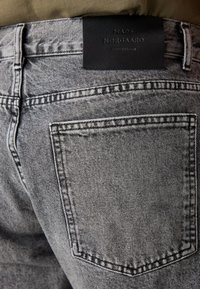 Jean en denim gris avec une finition texturée, présentant une poche arrière et une étiquette en cuir noir au niveau de la taille.
