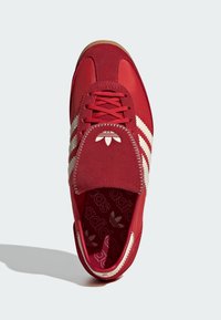 Červená atletická obuv s textilným zvrškom. Má biele bočné pruhy, polstrovaný jazyk a gumovú podrážku. Vo vnútri je vzor loga.