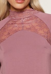 Čipkasti top u nježnoj boji mauve, s visokim ovratnikom i prozirnim čipkastim panelom preko ramena. Glatka tekstura tkanine s finim detaljima od čipke.