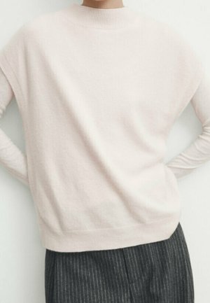 Person wearing a light beige turtleneck sweater and dark gray pinstripe pants with hands resting on hips.