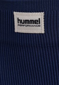 Hvid stoflabel med sort "hummel PERFORMANCE" tekst syet på ribbet marineblåt tekstil.