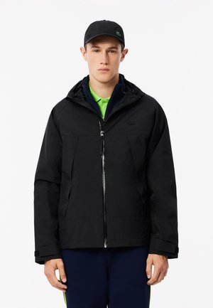 Zwarte waterdichte jas met een capuchon, voorzien van een ritssluiting aan de voorkant, twee grote zakken en verstelbare manchetten. Draagt over een felgroen shirt.