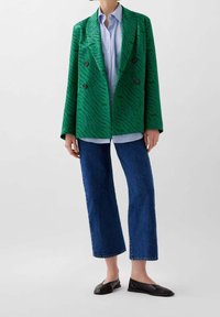 Blazer verde de doble botonadura con un patrón de rayas texturizadas, combinado con una camisa de rayas azul claro, jeans de pierna ancha oscuros y bailarinas negras.