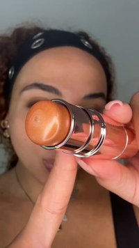 Person hält einen bronzefarbenen Make-up-Stift nahe an der Kamera, mit einem schwarzen Stirnband und lockigem Haar im Hintergrund.