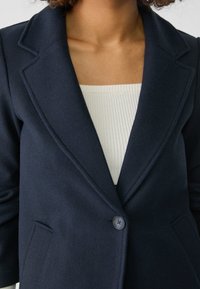 Marineblå blazer i uldblanding med hakkede revers, enkelt knaplukning og to forlommer; har en blød tekstur og skræddersyet pasform.