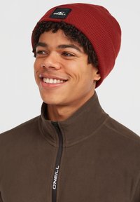 Gorro acanalado rojo con un parche cuadrado negro, usado con una chaqueta de polar marrón que cuenta con cremallera y cuello alto.