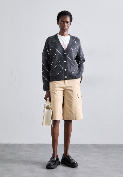 Og�ļu krāsa kardigāns ar baltiem pērļu akcentiem, ar pogu aizdari un divām kabatām, kopā ar bēša krāsas kravas šortiem un melniem masīviem loaferiem.