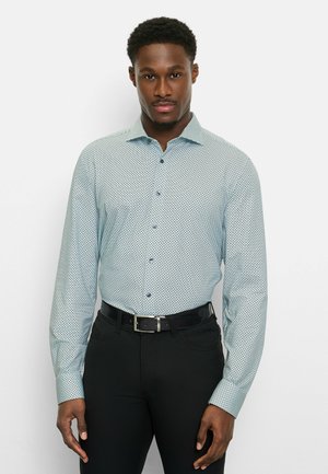 Mann trägt hellblau gemustertes Hemd mit langen Ärmeln, in schwarze Hose mit schwarzem Gürtel gesteckt, steht vor weißem Hintergrund.