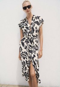 Robe à motifs floraux noir et blanc avec un col, des manches courtes, un devant boutonné, une taille ceinturée et une fente sur le côté, fabriquée en tissu léger.