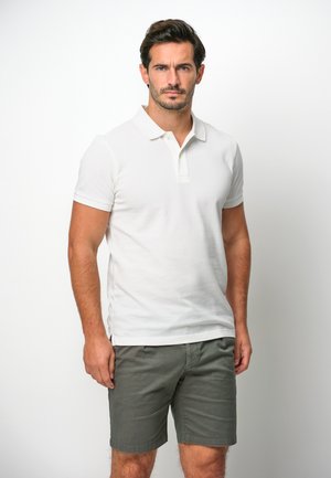 Hvid polo skjorte med korte ærmer, krave og sidelæg, parret med mørkegrå shorts, sat mod en ensfarvet baggrund.