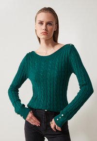 Teal cable-knit sweater with a wide neckline and ribbed cuffs. Gold-button accents on shoulders. Worn with black high-waisted jeans.