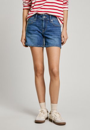 Mujer con pantalones cortos de mezclilla azules, zapatillas blancas con calcetas beige y una camiseta de manga larga a rayas rojas y blancas, de pie con los brazos relajados.