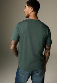 Tricou din bumbac verde pădure, cu mânecă scurtă, guler rotund și croială lejeră, purtat împreună cu blugi din denim albastru.