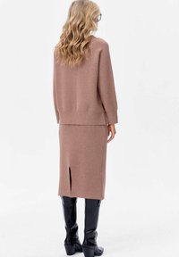 Sweter i spódnica w kolorze jasnobrązowym wykonane na drutach, z bocznym rozcięciem. Modelka nosi czarne kozaki za kolano, podkreślając gładkie tekstury i prosty design.