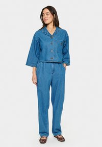 Skrajšana jeans jakna s ovratnikom, gumbi in žepom na prsih, v kombinaciji s širokimi jeans hlačami. Obe sta v srednjem modrem pranju.