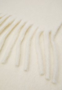 Kreemjas tekiks, mis on valmistatud pehmest ja karvase materjalist, ühe serva ääres on särgid, demonstreerides sujuvat tekstuuri ja sooja väljanägemist.
