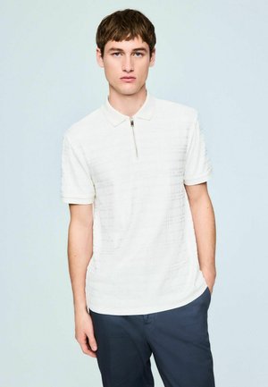 Jauns vīrietis baltā īsroku polo krekliņā ar priekšējo rāvējslēdzēju un tumšām biksēm, stāvot pret gaišu fonu.