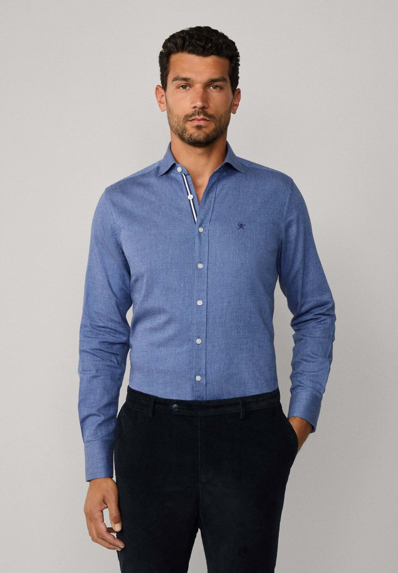 Chemise bleue à boutons dans un tissu texturé. Dispose d'un col intérieur à rayures contrastées et d'un petit logo brodé sur la poitrine. Associée à un pantalon noir.
